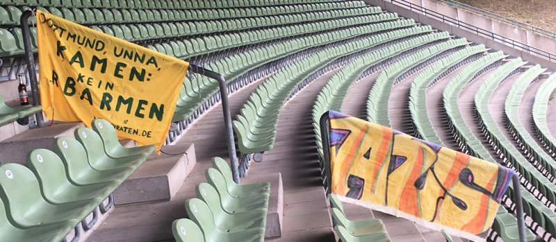 2 Banner auf der Tribüne des Stadion Hohenhorst. Links: »Dortmund. Unna. Kamen. Kein Erbarmen. vestpiraten.de«; rechts: AuswärZSieg in bunt. Im Hintergrund die leeren Plätze der Sitztribüne